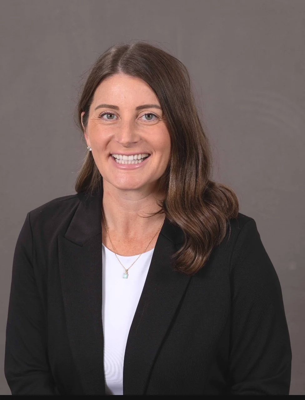 Professional woman with long brown hair wearing a black blazer and white blouse, smiling against a plain gray background.