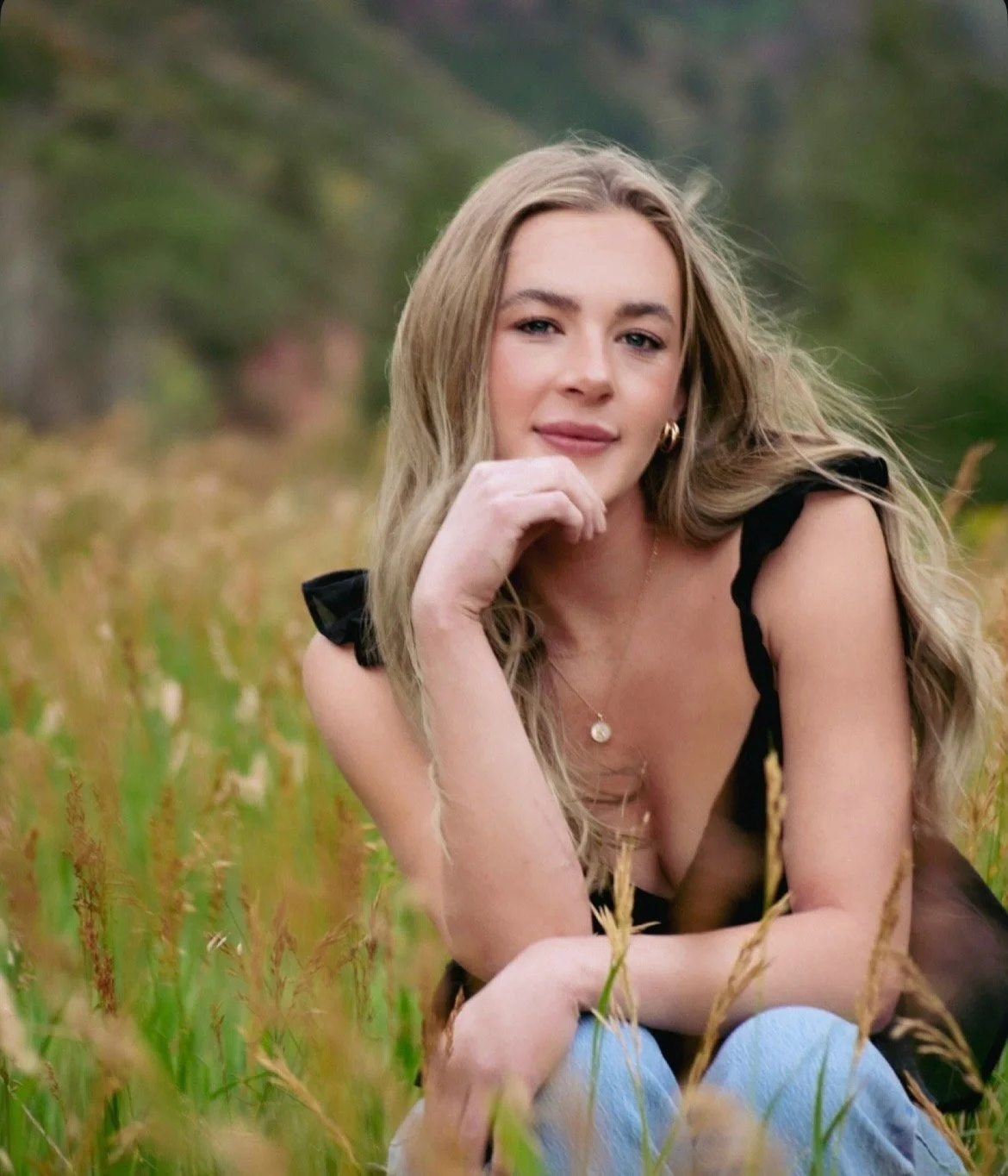 A young woman with blonde hair, wearing a black top with ruffled sleeves, is sitting in a grassy field in Colorado with trees in the background. She is resting her chin on her hand and looking at the camera.