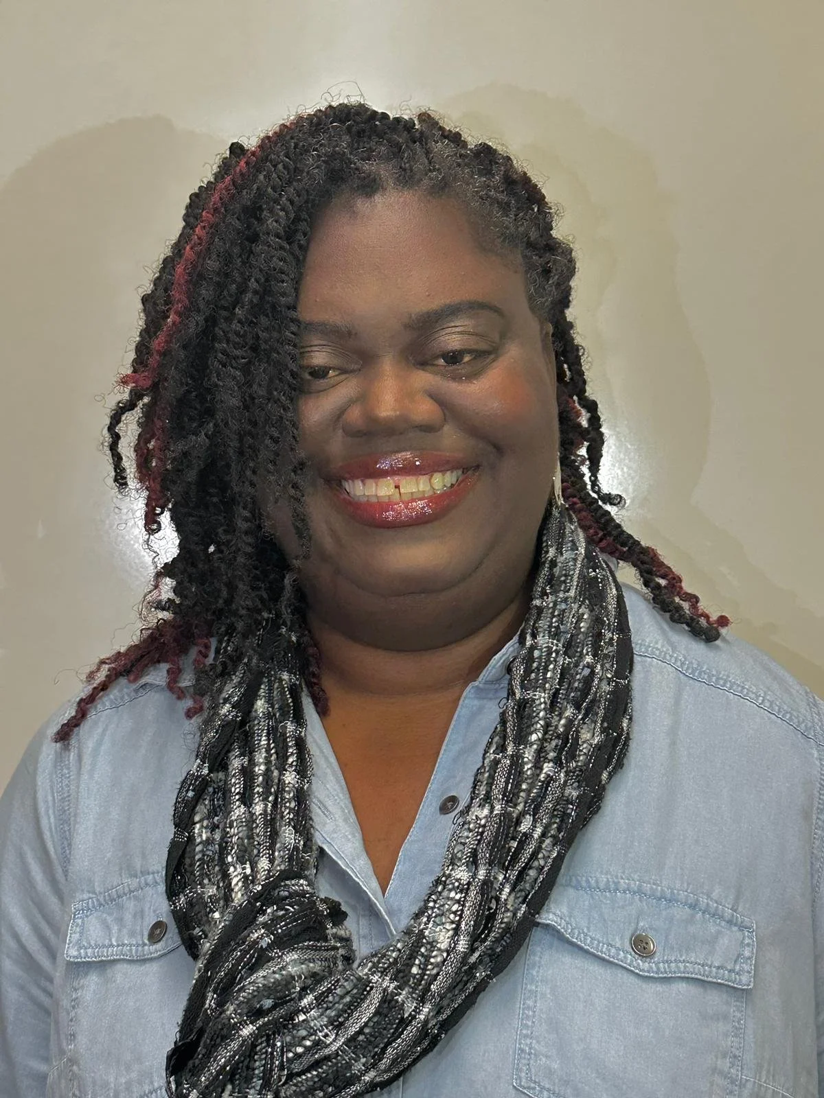 A woman with styled black and red twist braids, wearing a light denim shirt and a black, white, and silver scarf, smiling in front of a plain wall.