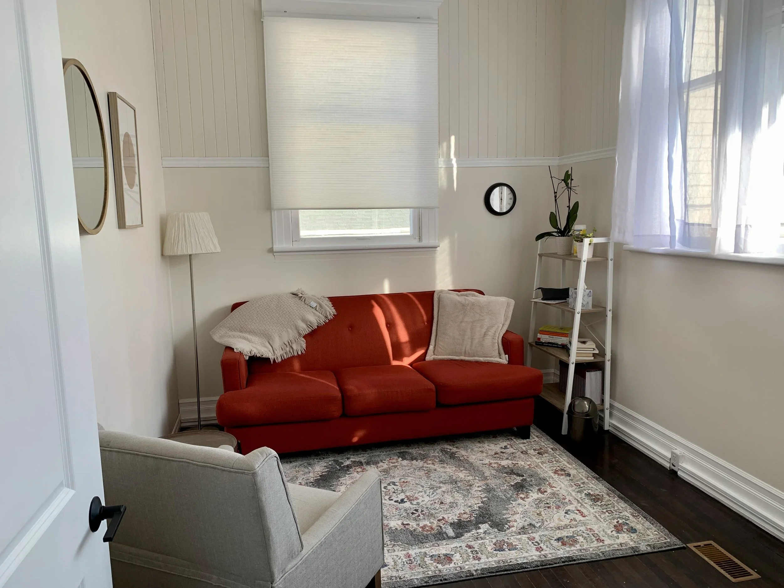 Living room with a red sofa, beige armchair, white bookshelf, plants, window with curtains, wall clock, and framed artwork, illuminated by natural light.