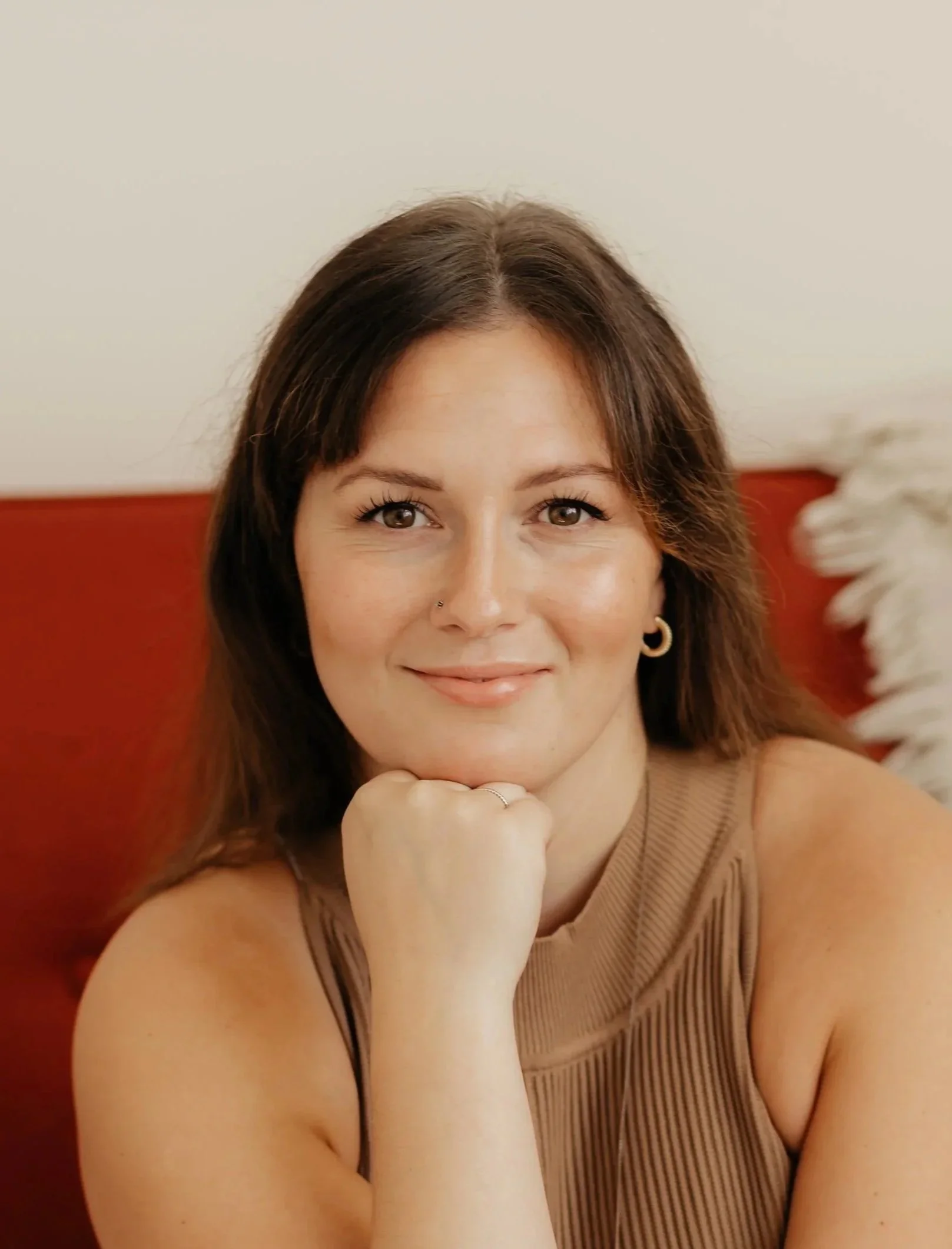 A woman with long wavy brown hair and a slight smile, wearing a brown top, against a light background.