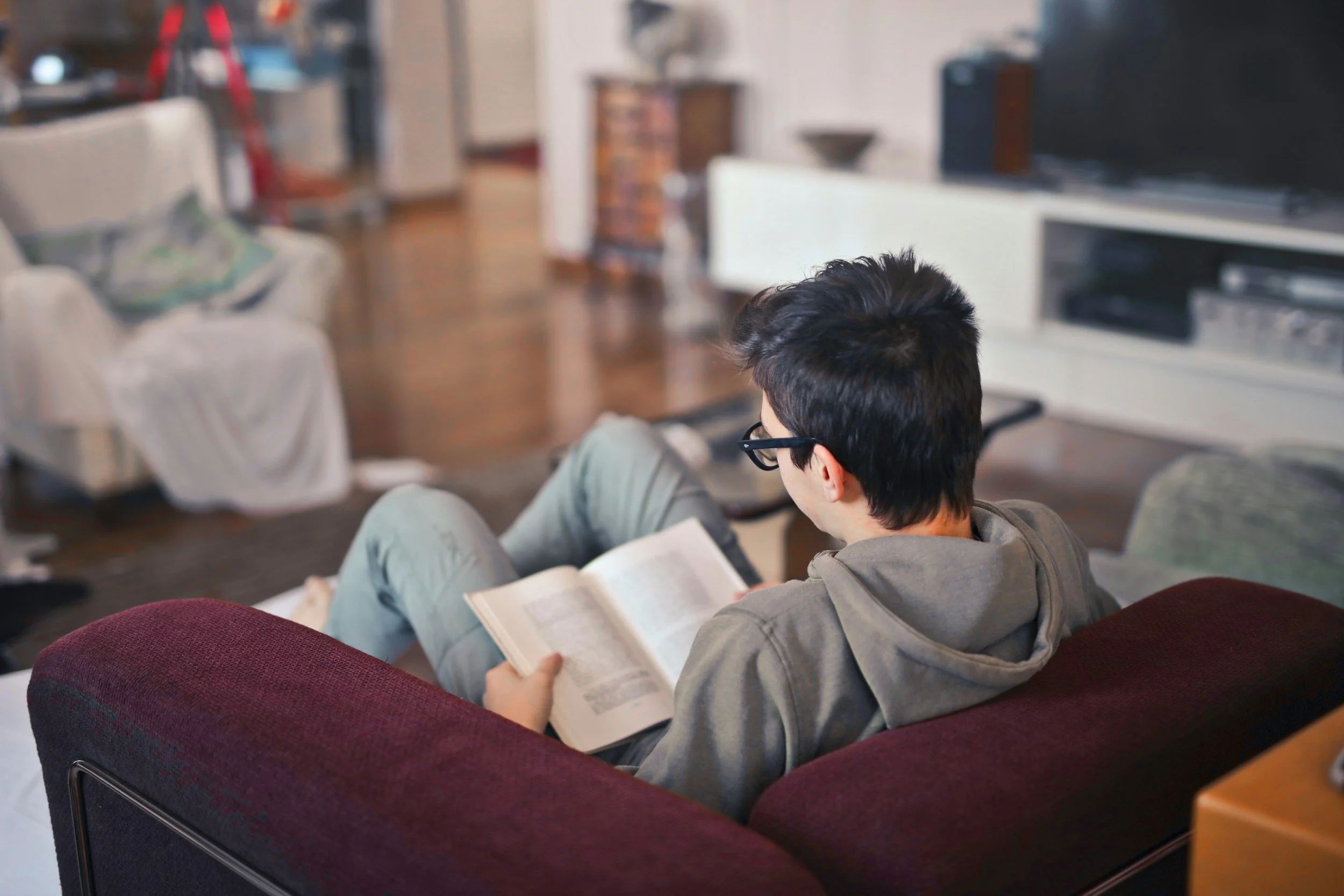 A person with black hair and glasses, sitting on a maroon couch in a living room, reading a book.