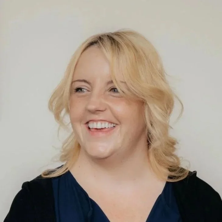 A woman with blonde, shoulder-length hair smiling and looking to the side, wearing a dark top, standing against a plain, light-colored background.