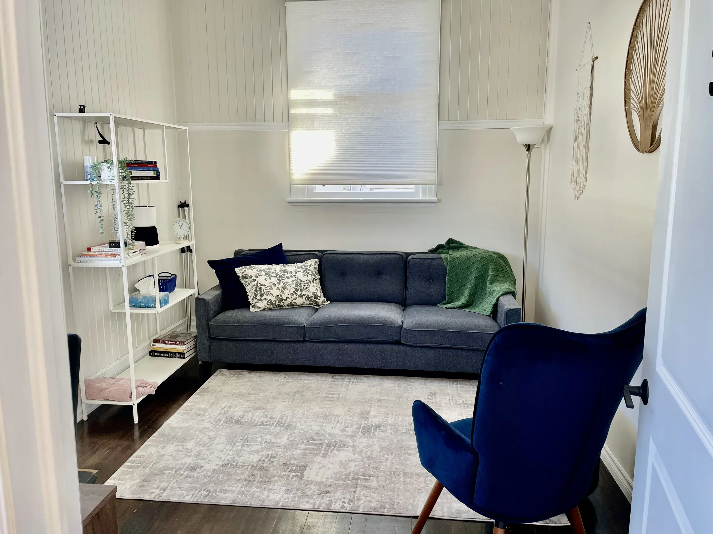 A cozy living room with a dark blue couch, a tall white bookshelf on the left, a blue armchair in the foreground, a window with a white blind, a beige rug, and a floor lamp on the right, with decorative wall hangings.
