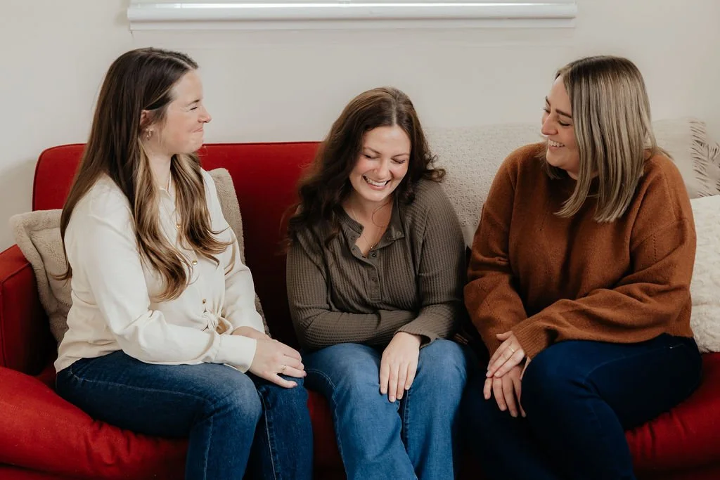 Three women sitting on a red couch, laughing and talking with each other.