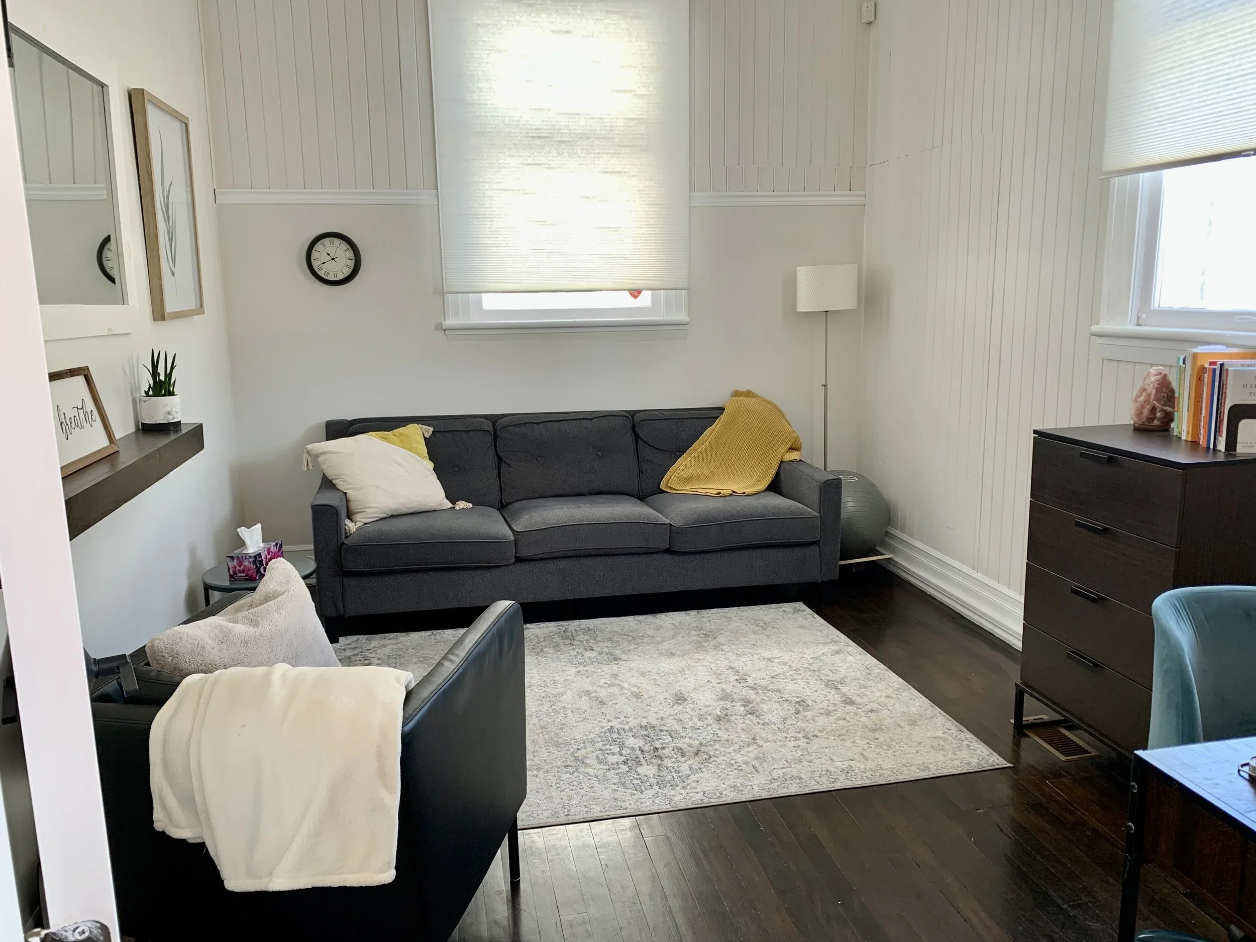 Living room with dark gray sofa, black armchair, white walls, wooden floor, decorative wall art, clock, window with blinds, bookshelf, and side table.