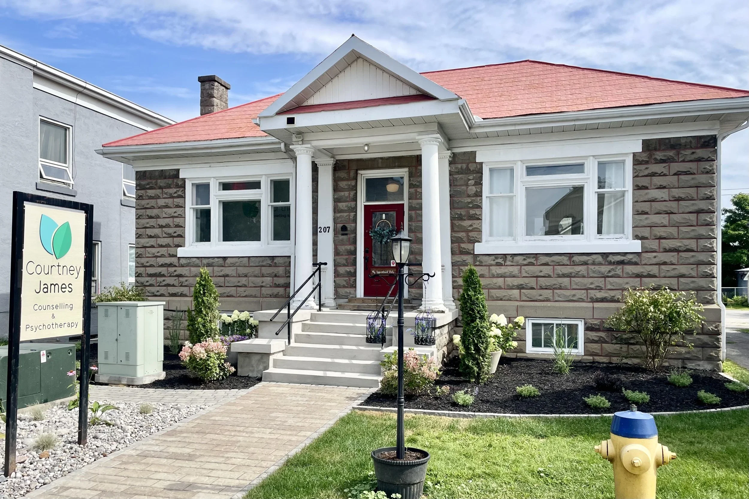 A brick house with white columns and a red roof, a front porch with steps, and a landscaped yard with flowers and shrubs. There is a sign that reads 'Courtney James Counseling & Psychotherapy' and a black lamppost with hanging flower baskets.