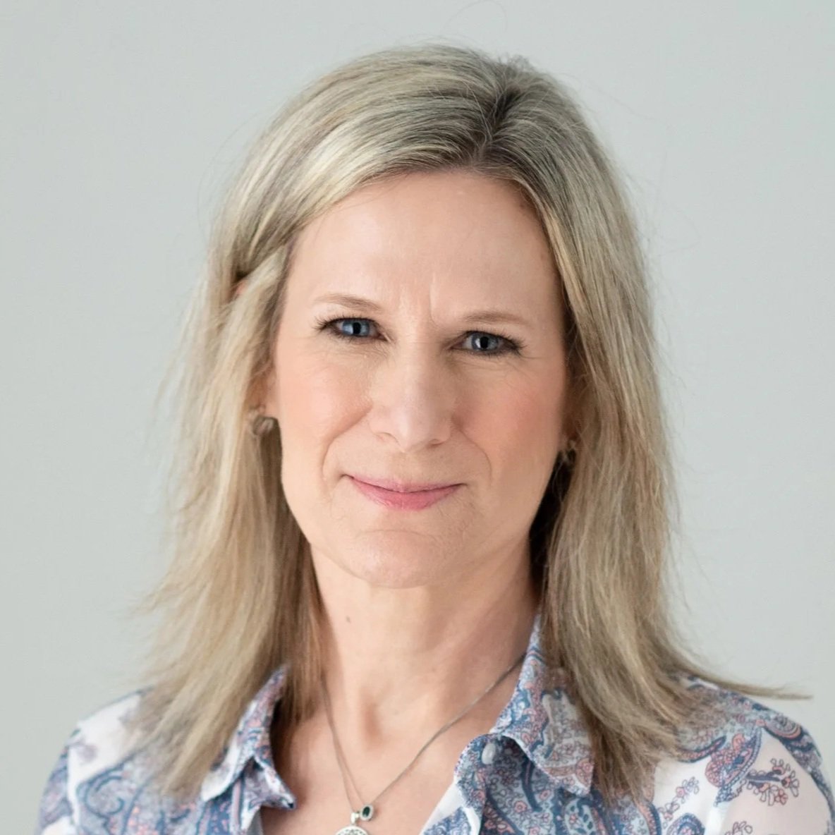 A woman with blonde hair, light skin, and blue eyes smiling slightly at the camera, wearing a patterned shirt and a necklace, against a plain light background.