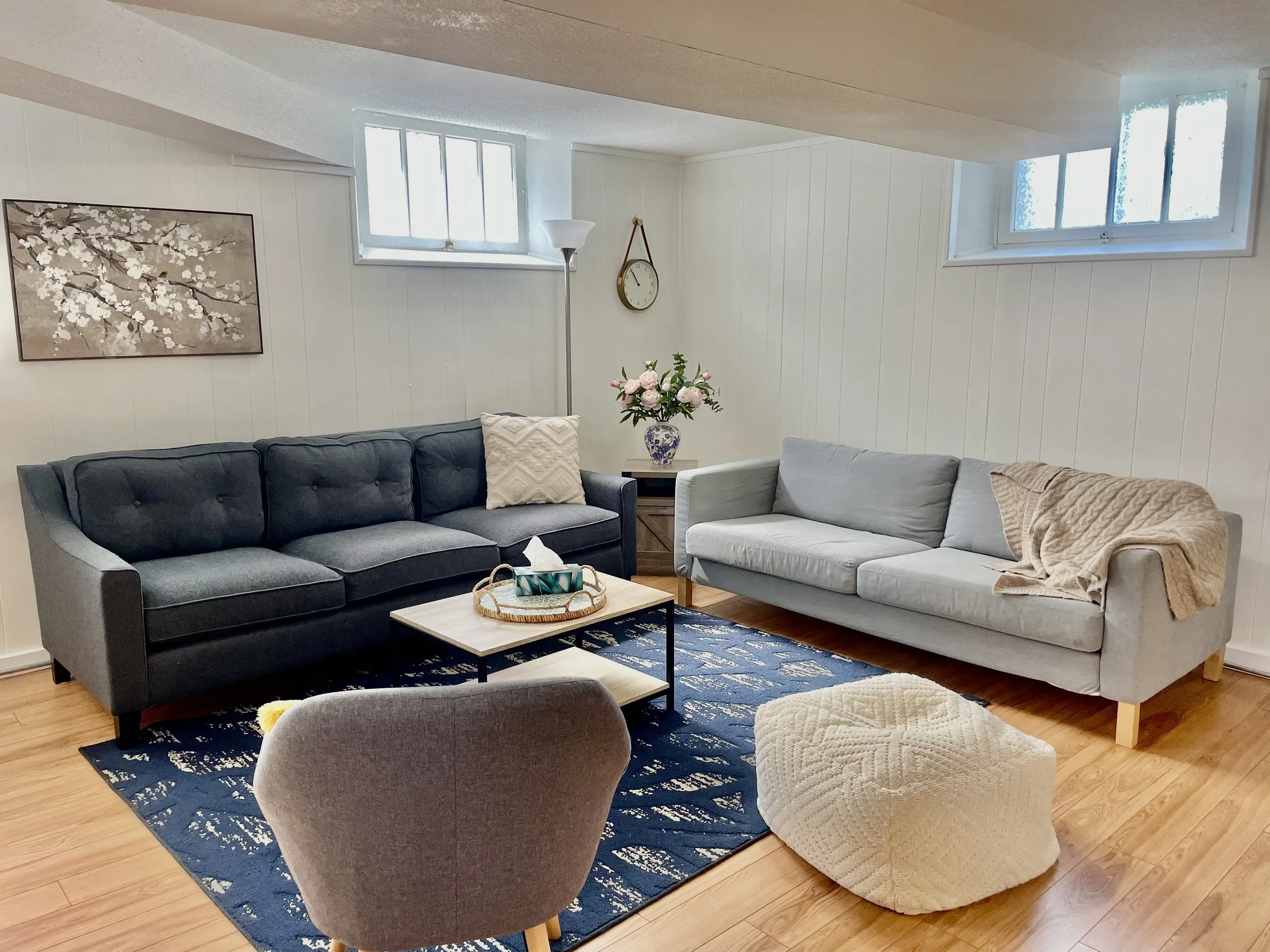 Living room with two sofas, a coffee table, a patterned blue rug, an armchair, and a textured pouf. The room has wood flooring, white paneled walls, two windows, a flower vase, a wall clock, a painting, and a floor lamp.
