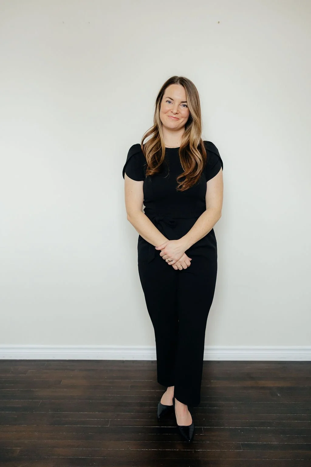 A woman with long, wavy brown hair wearing a black outfit and black high heels, standing against a plain white wall with her hands clasped in front of her, smiling.