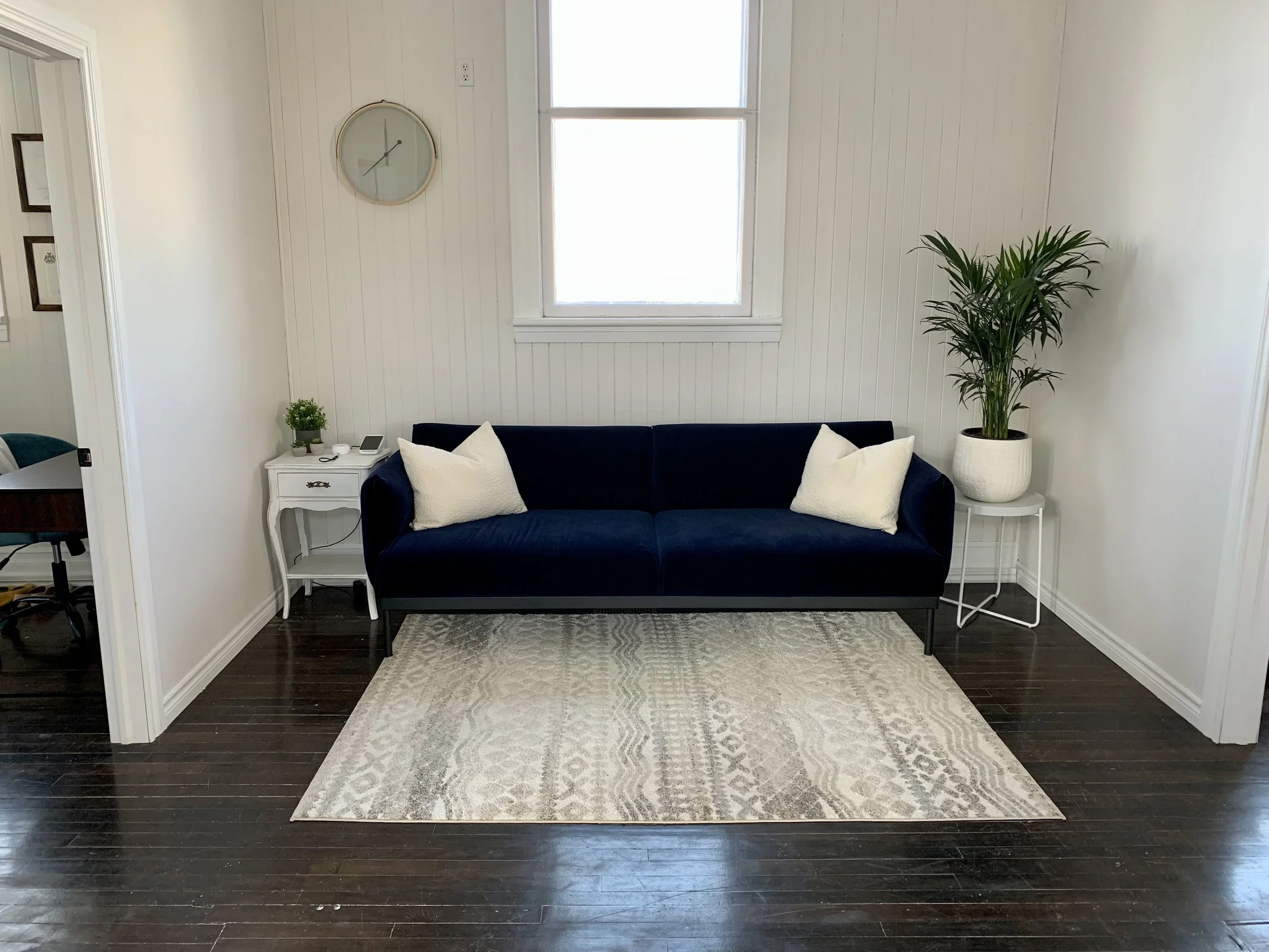 Living room with a navy blue sofa, white pillows, a white side table with a lamp and plant, and a large potted plant on a small white table, with a window and wall clock in the background.