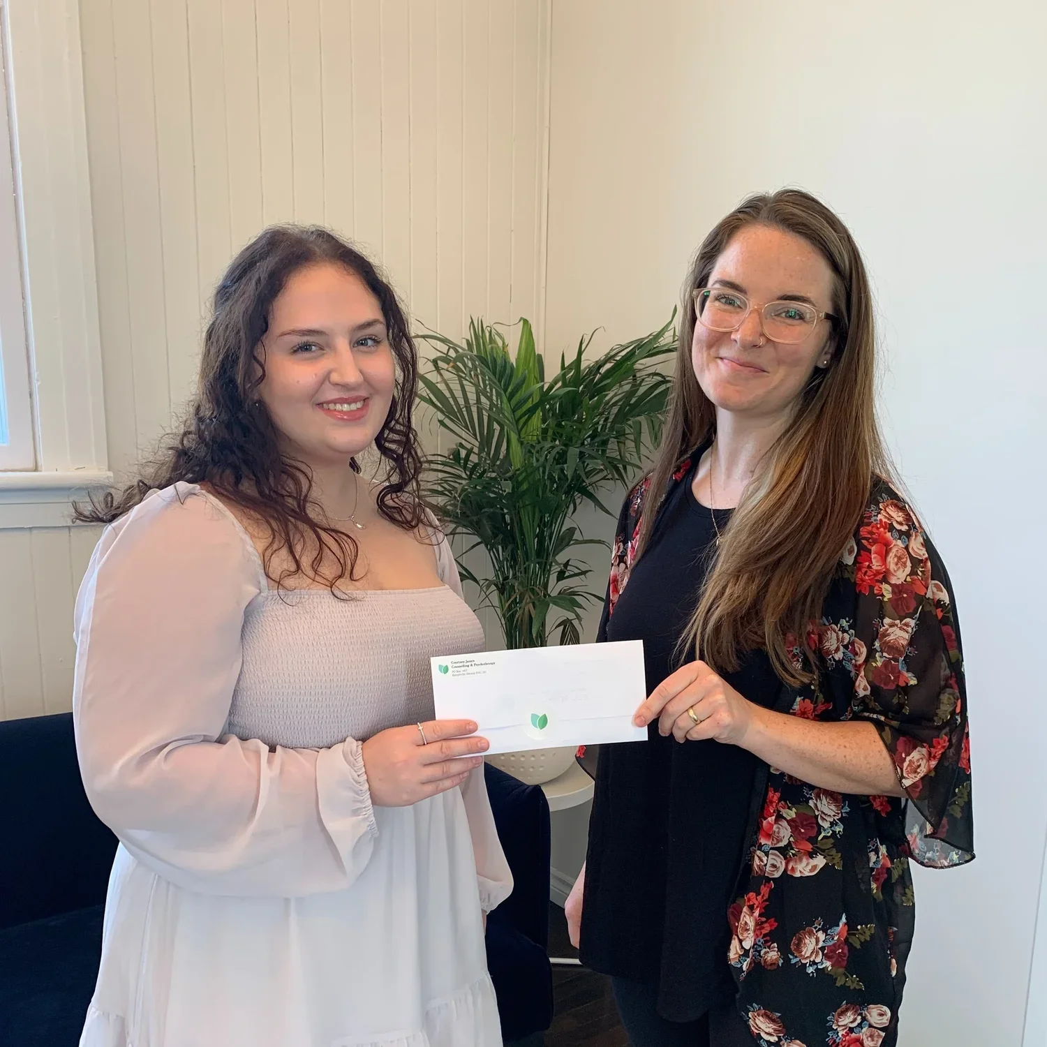 Two women standing inside a room, one holding a check, smiling at the camera. The woman on the left has curly brown hair, wearing a beige dress. The woman on the right has long straight brown hair, wearing glasses and a black floral blouse. A potted plant is visible behind them.
