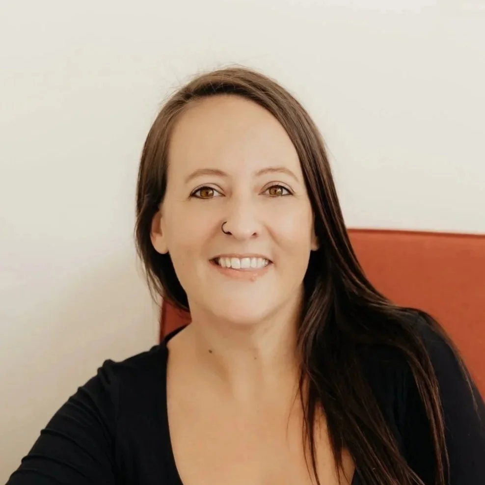 A woman with long brown hair and a nose ring smiling at the camera, sitting indoors with a white wall and red headboard in the background.
