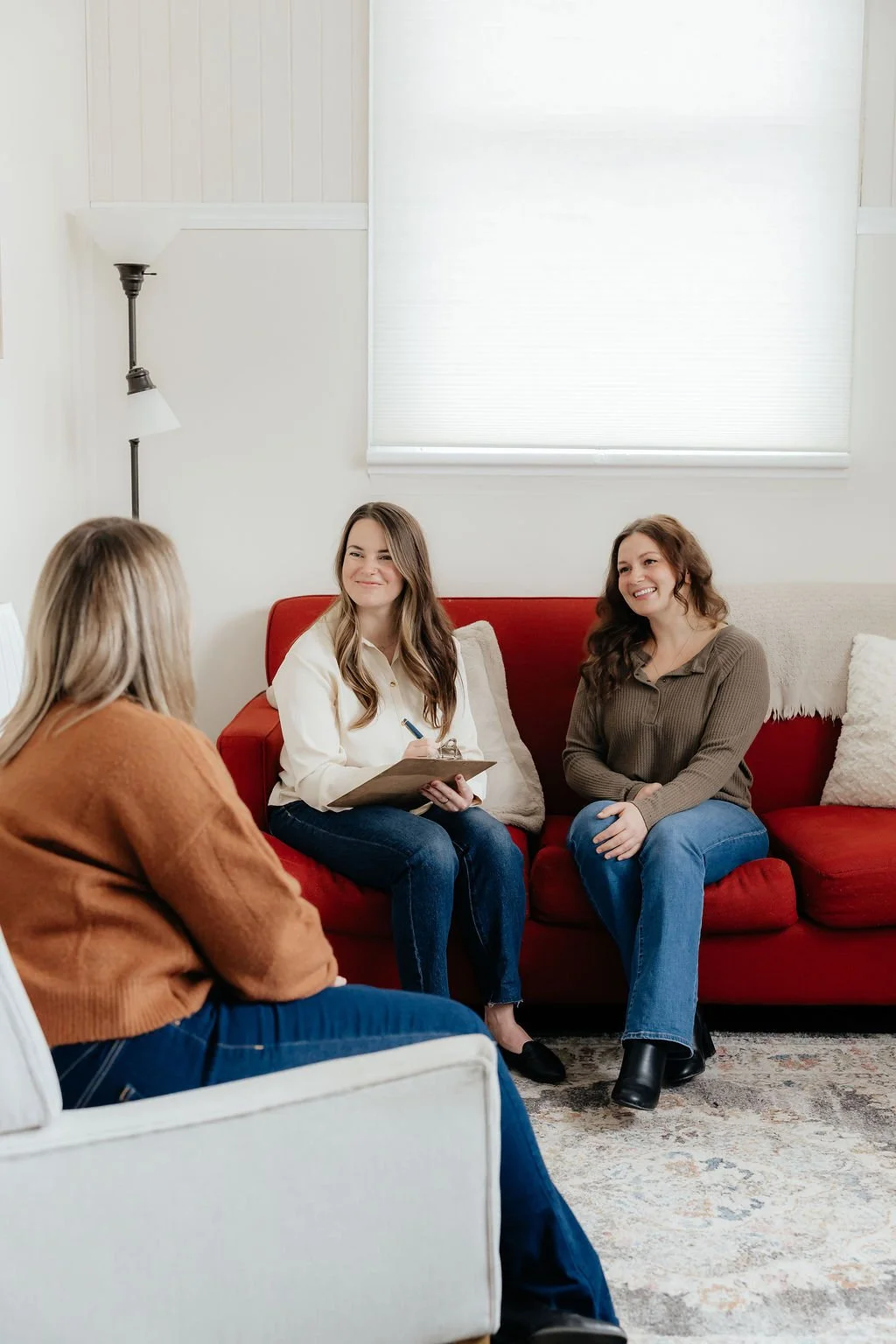 Three women sitting on a red sofa and a white chair in a bright room, having a conversation, with one woman holding a clipboard.