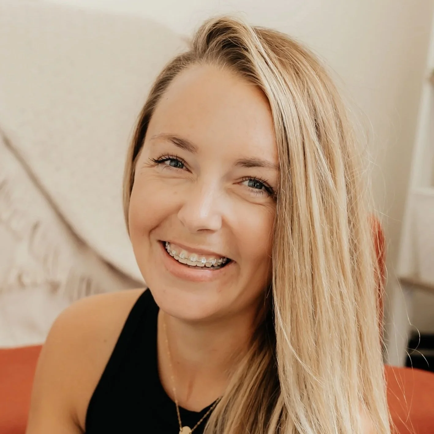A woman with blonde hair smiling and showing braces, wearing a black top, sitting on an orange or red couch, with a cream-colored background.