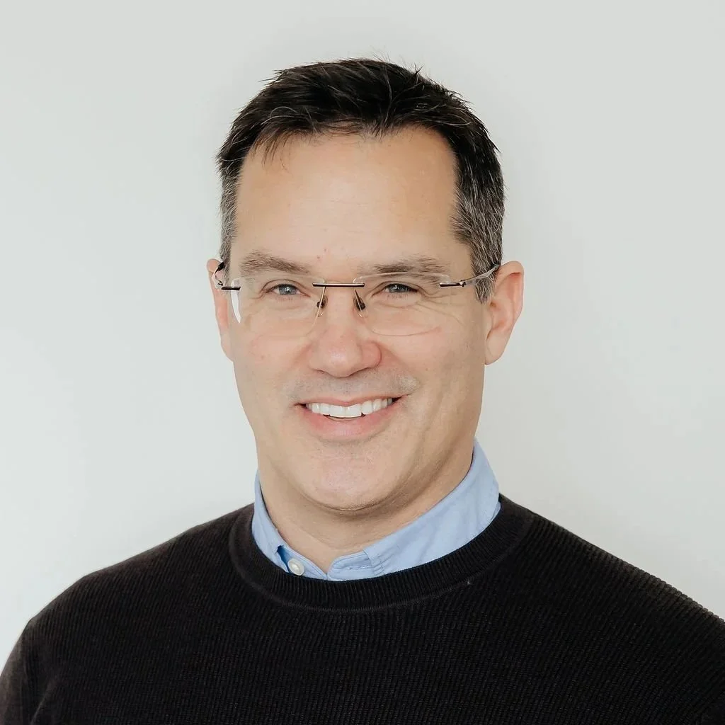 A man with short dark hair, wearing glasses, a light blue collared shirt, and a black sweater, smiling at the camera against a plain white background.