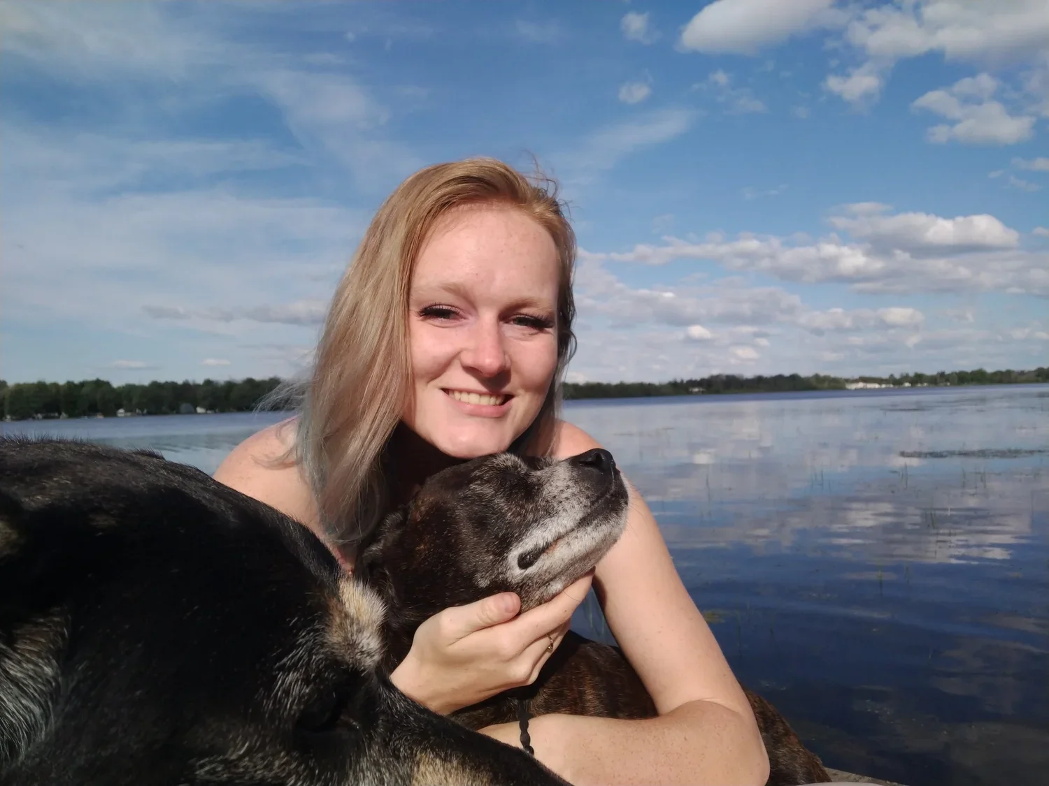 A young woman with long blonde hair smiling and hugging a large brindle dog near a lake with a partly cloudy sky.
