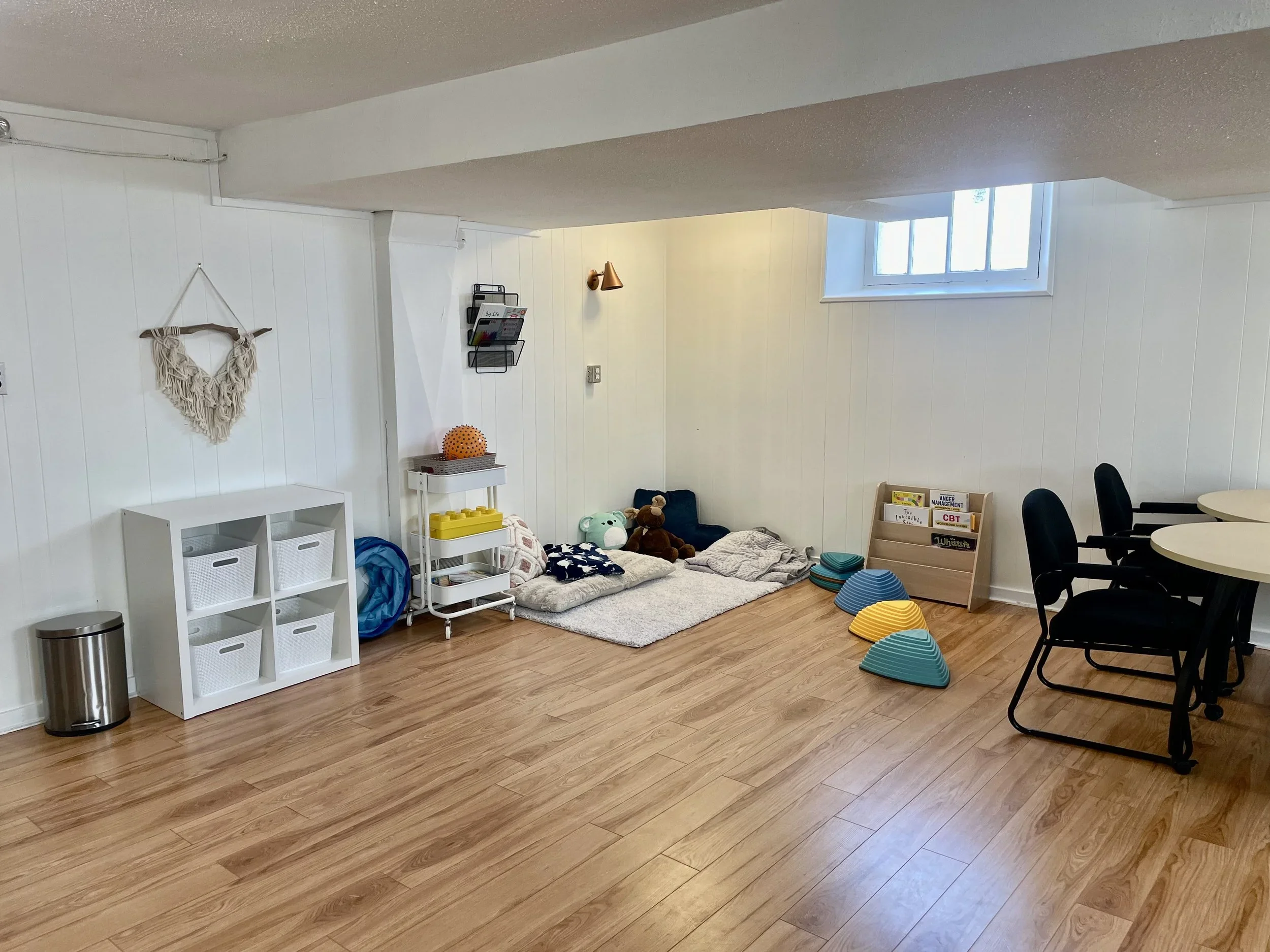 Children's play area with cushions, stuffed animals, and books on a wooden floor, with a small window and white paneled walls.