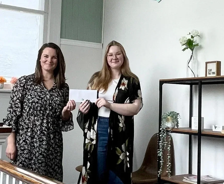 Two women standing indoors, smiling, one holding a white envelope or card.