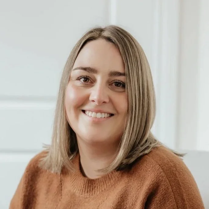 A young woman with shoulder-length blonde hair smiling at the camera, wearing a brown sweater in a bright room with white walls.