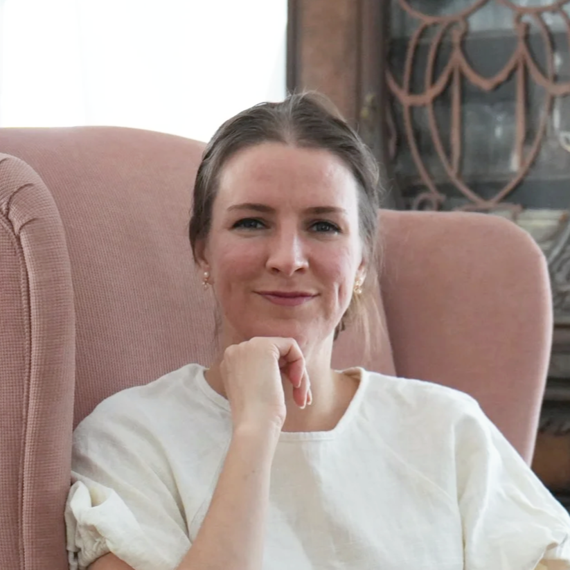 A woman sitting on a pink armchair, smiling with her hand resting on her cheek. She has dark hair pulled back, light skin, and is wearing a white shirt with pearl earrings. There is a decorative piece on the wall behind her.