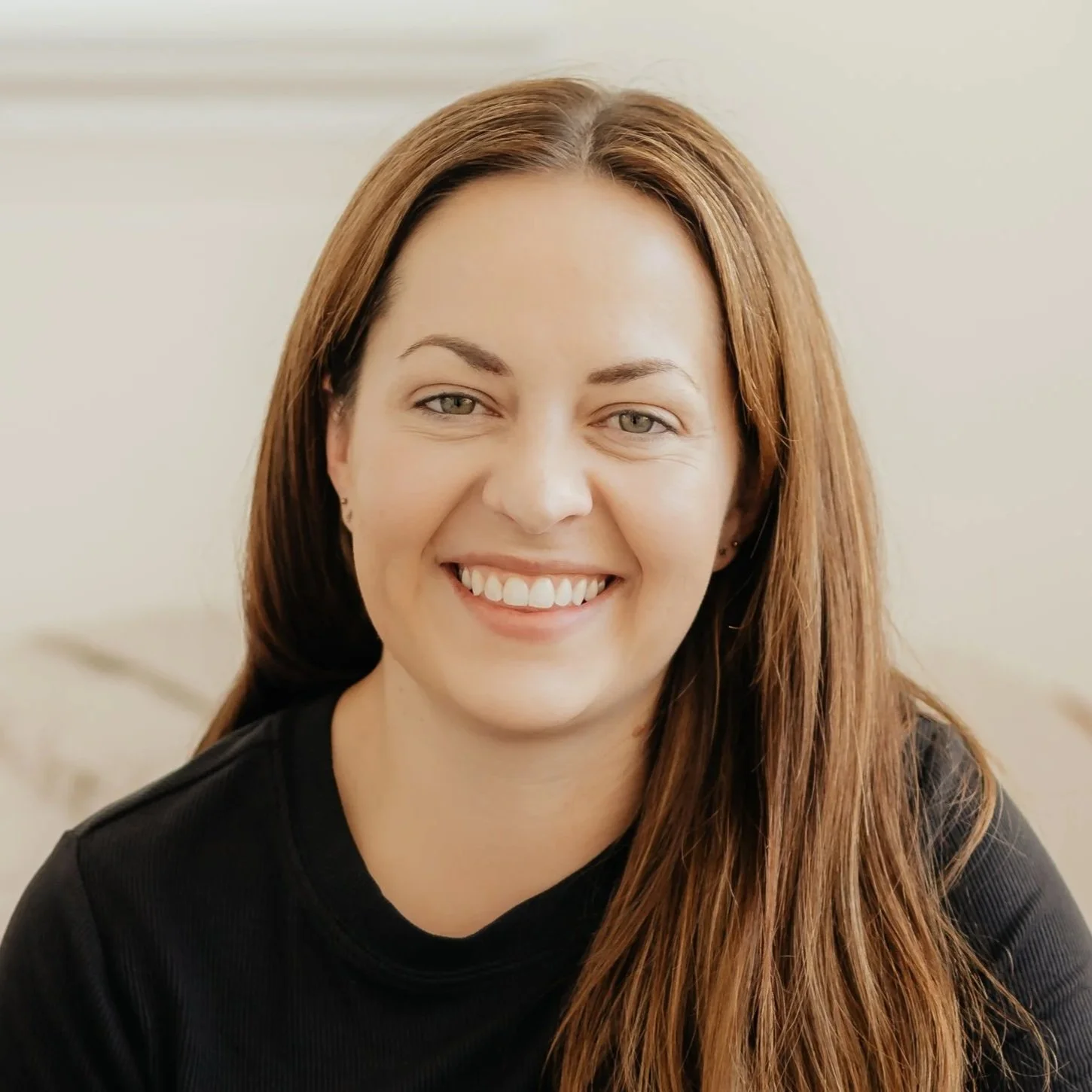 Close-up of smiling woman with long auburn hair and light makeup, wearing a black top, in a neutral background setting.