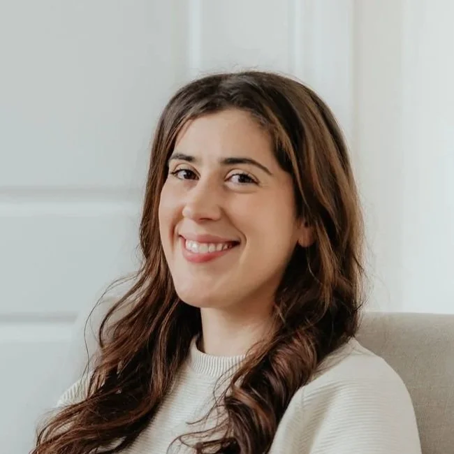A young woman with long brown hair sitting on a beige chair, smiling at the camera, in a room with white walls.