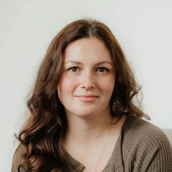 A woman with long wavy brown hair and a slight smile, wearing a brown top, against a light background.