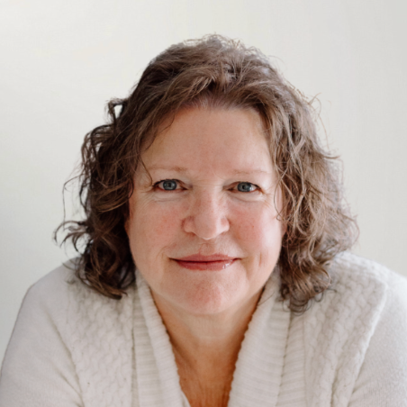 Close-up portrait of a woman with curly brown hair, blue eyes, and a slight smile, wearing a white sweater, against a plain light background.