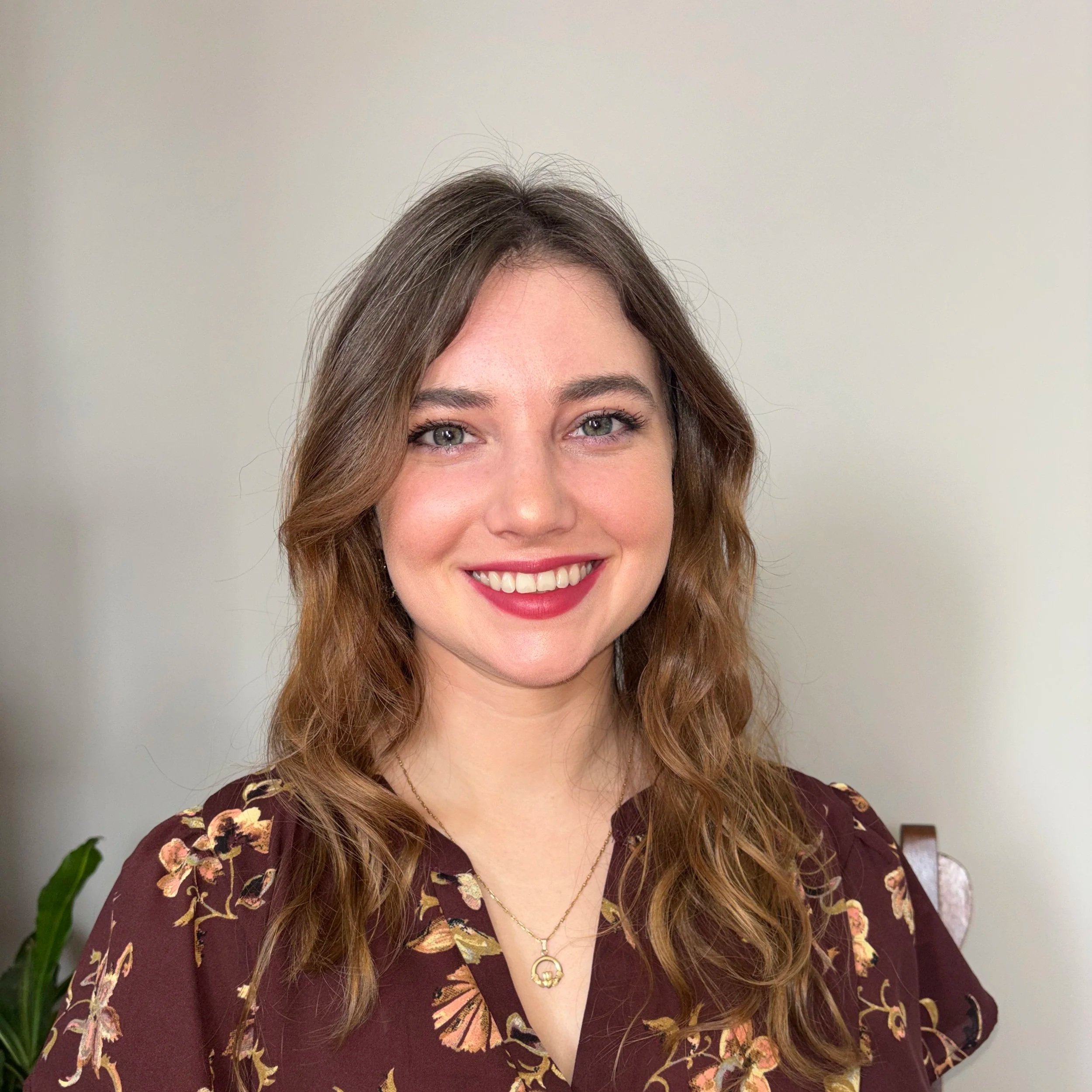 A young woman with wavy brown hair and blue eyes, smiling, wearing a maroon floral top and a gold necklace, standing in front of a plain background.
