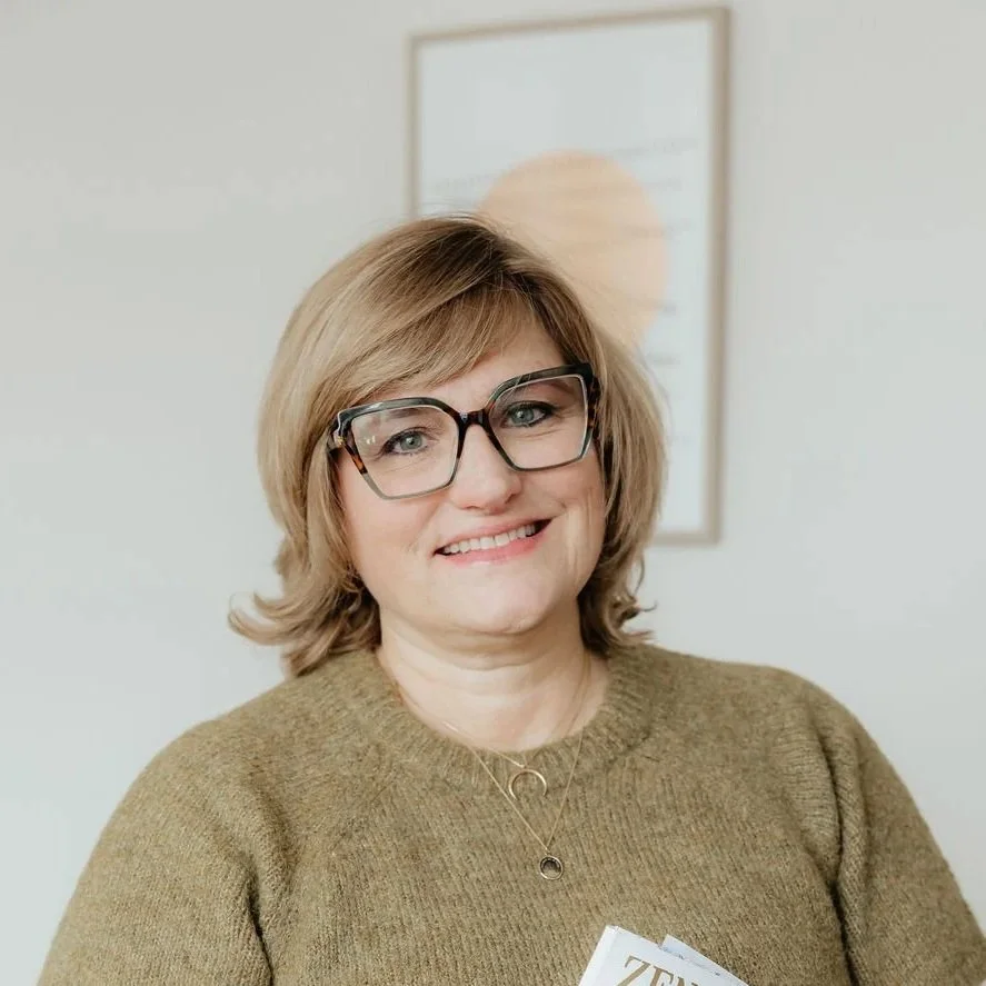 A smiling woman with shoulder-length blond hair, wearing large black glasses and a brown sweater. She has layered necklaces and is standing in front of a light-colored wall with a framed certificate or document.