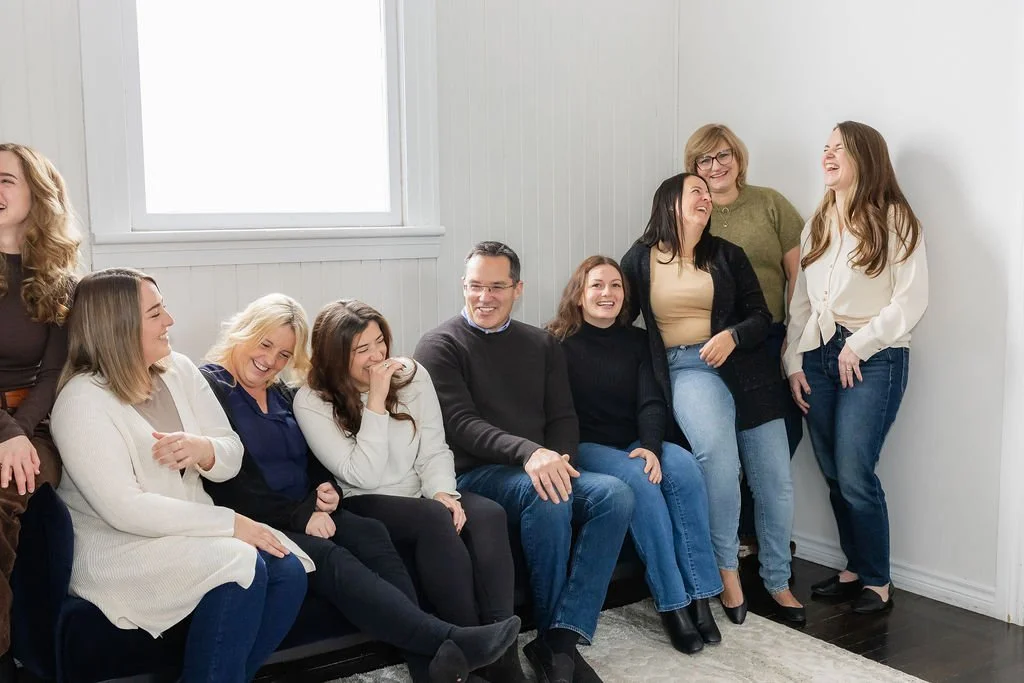 Group of smiling women and one man sitting and standing in a light-filled room, sharing a laugh.