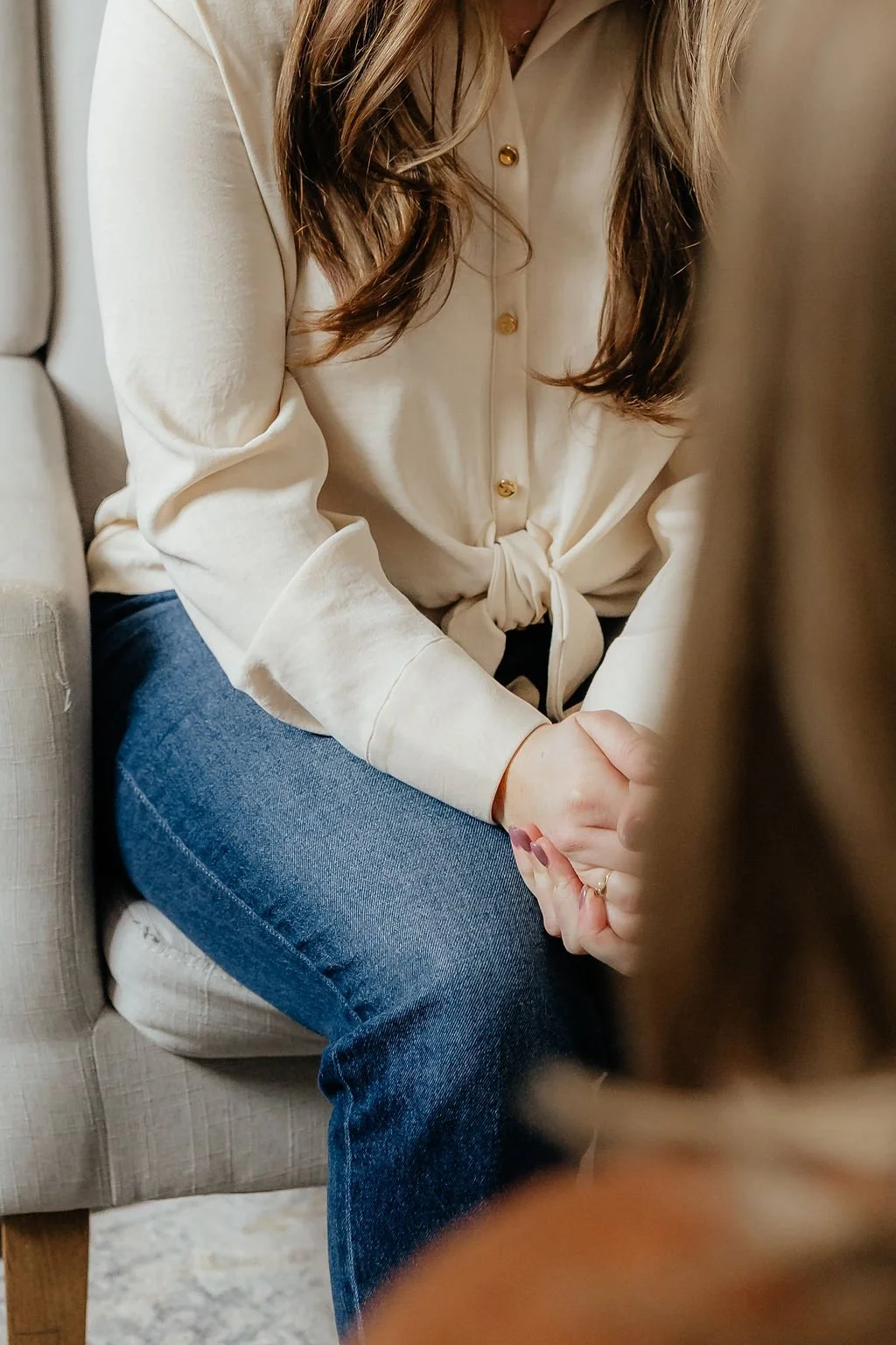 Close-up of a woman wearing a cream-colored shirt with gold buttons and blue jeans, sitting on a beige armchair, clasping her hands together.