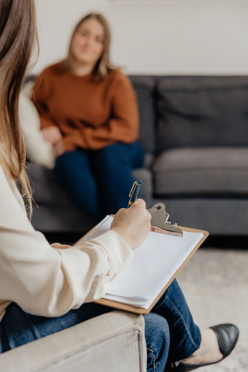 Therapist writing notes on a clipboard during a counseling session with a woman sitting on a sofa.