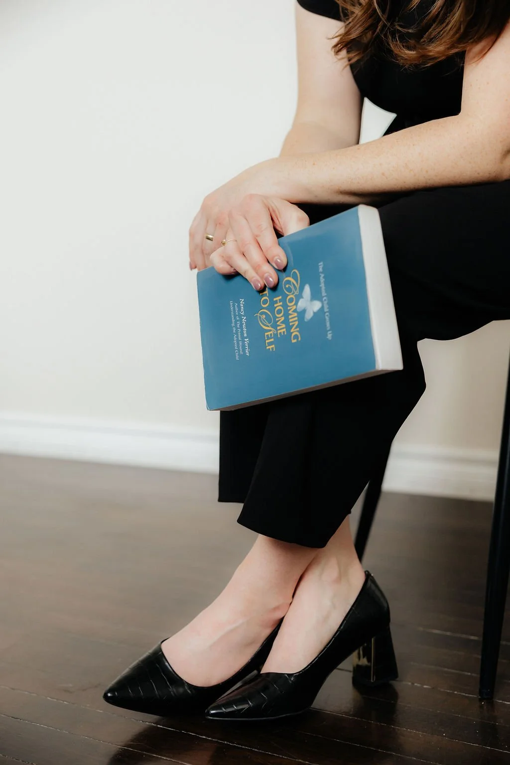 A woman sitting on a black chair, wearing black pants and black high heel shoes, holding a book titled "Coming Home to Joy".
