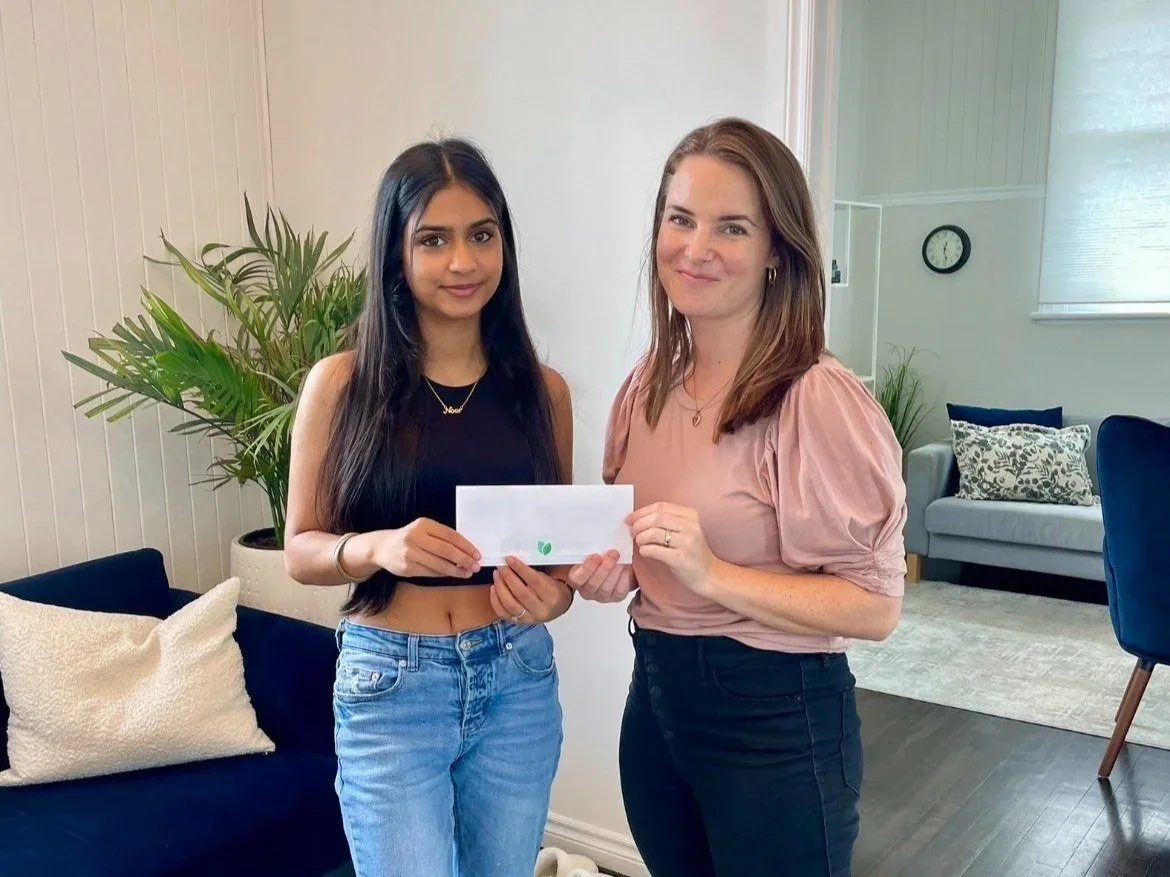 Two women standing indoors, holding an envelope together, with a living room background including a plant, a fireplace, and furniture.