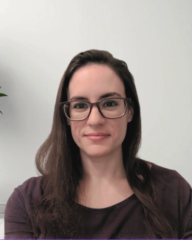 A woman with long brown hair and glasses smiling at the camera, wearing a dark-colored shirt, with a plain white wall in the background.