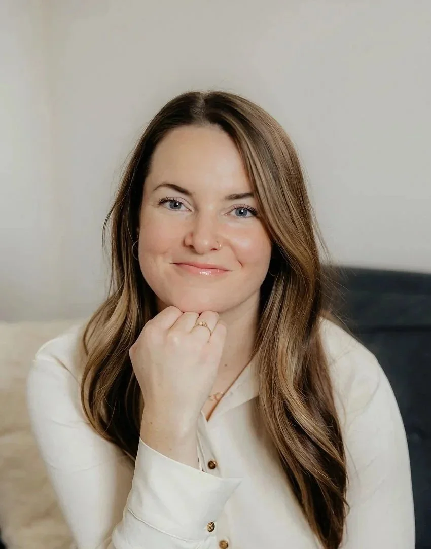 A woman with long brown hair and a nose ring, smiling and resting her head on her hand, wearing a light blue blouse in an indoor setting.