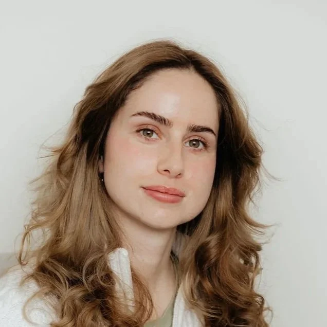 A young woman with wavy brown hair and fair skin smiling softly at the camera against a plain white background.