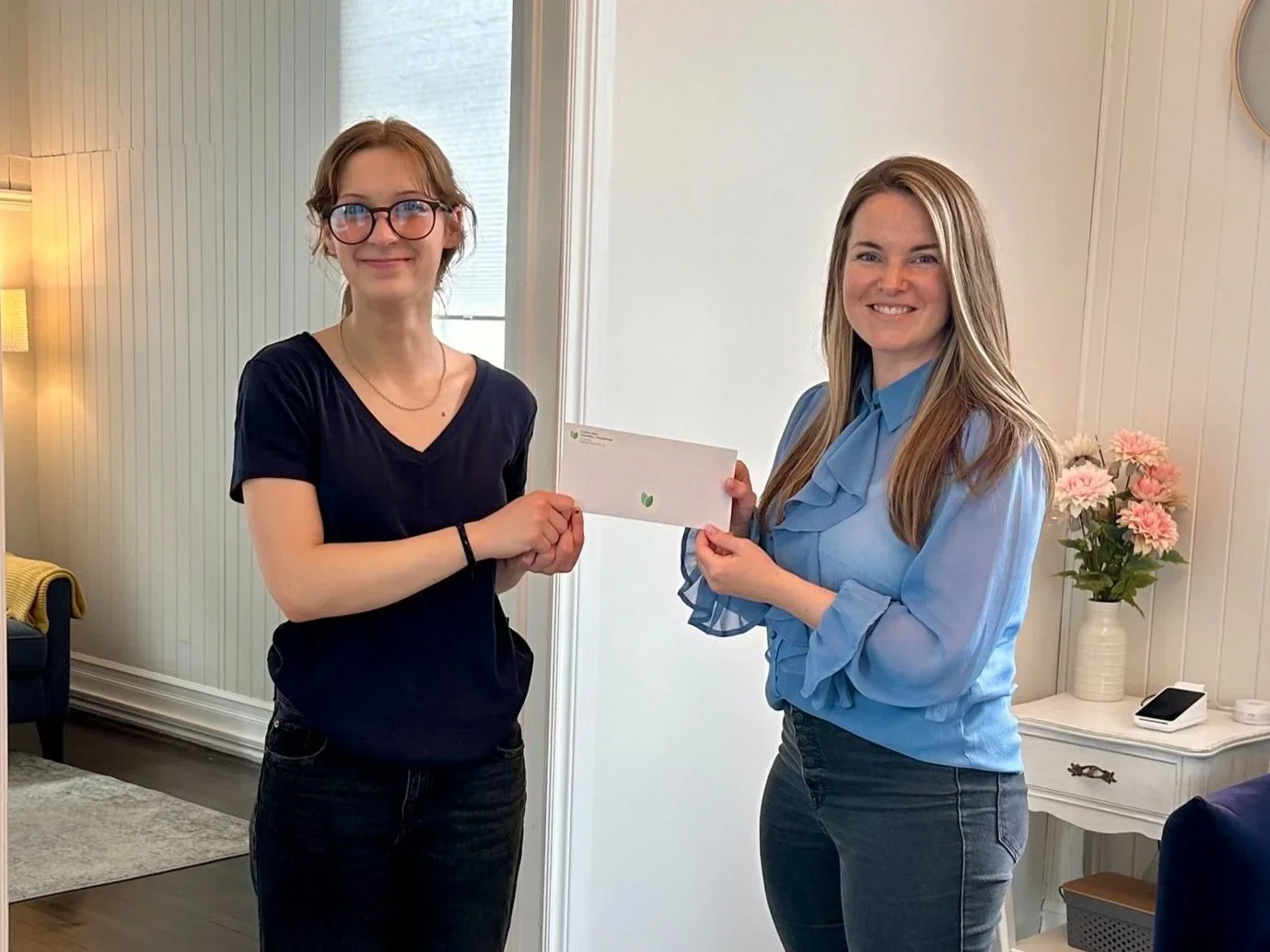Two women standing indoors, exchanging a white envelope, smiling at the camera, with a side table holding pink flowers and a phone in the background.