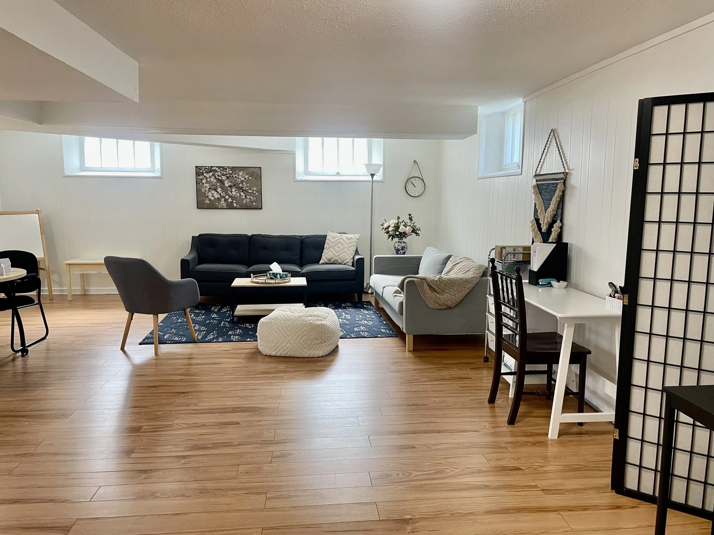Living room with hardwood floors, black and gray sofas, a coffee table, a white pouf, and a desk with a black chair, decorated with wall art, a clock, and a flower arrangement.
