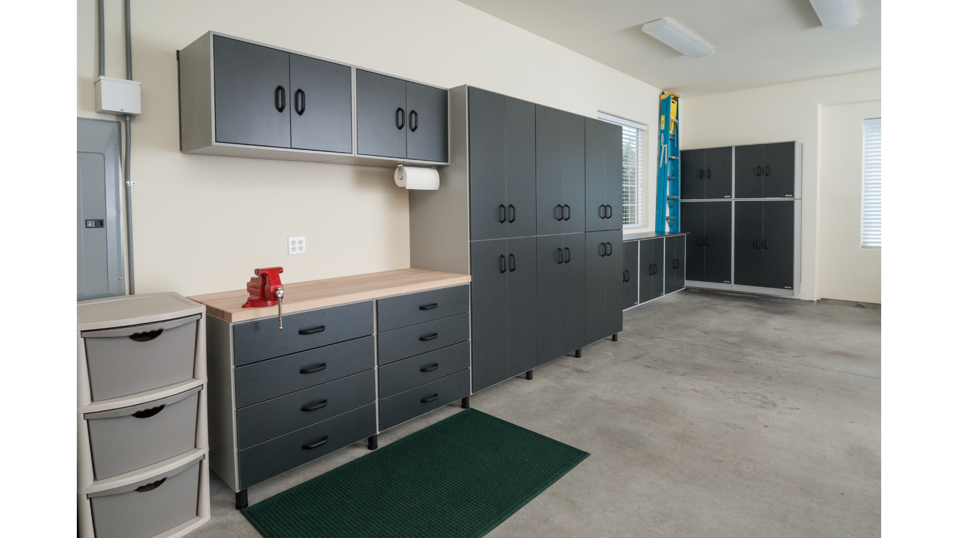 Garage with grey storage cabinets, a workbench, plastic drawers, a ladder, and windows with blinds.