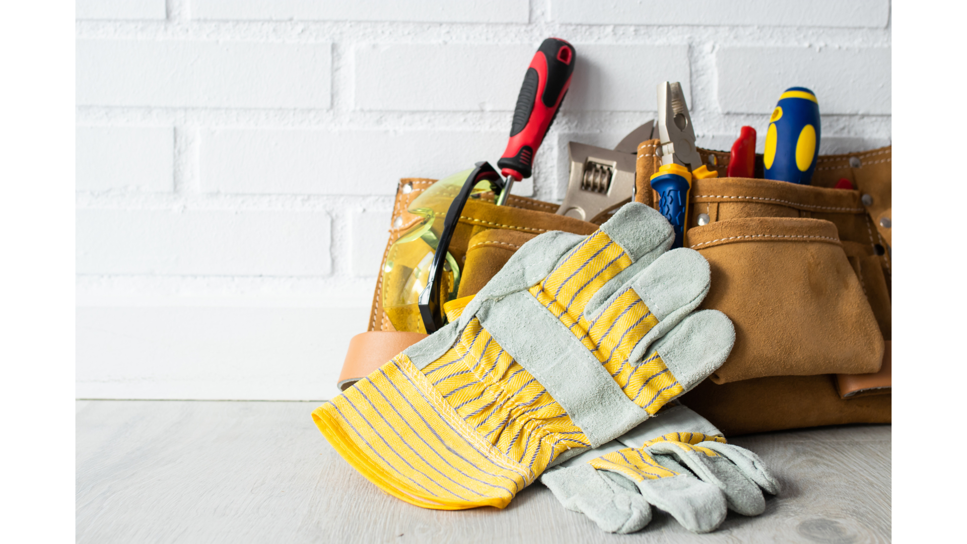A tool belt filled with various tools including a screwdriver, pliers, and a tape measure, along with work gloves and safety goggles, set against a white brick wall.