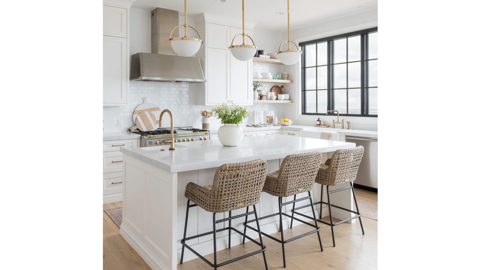 Bright, modern kitchen with white cabinetry, a large white island, woven bar stools, black-framed windows, and gold accents on fixtures and lighting.