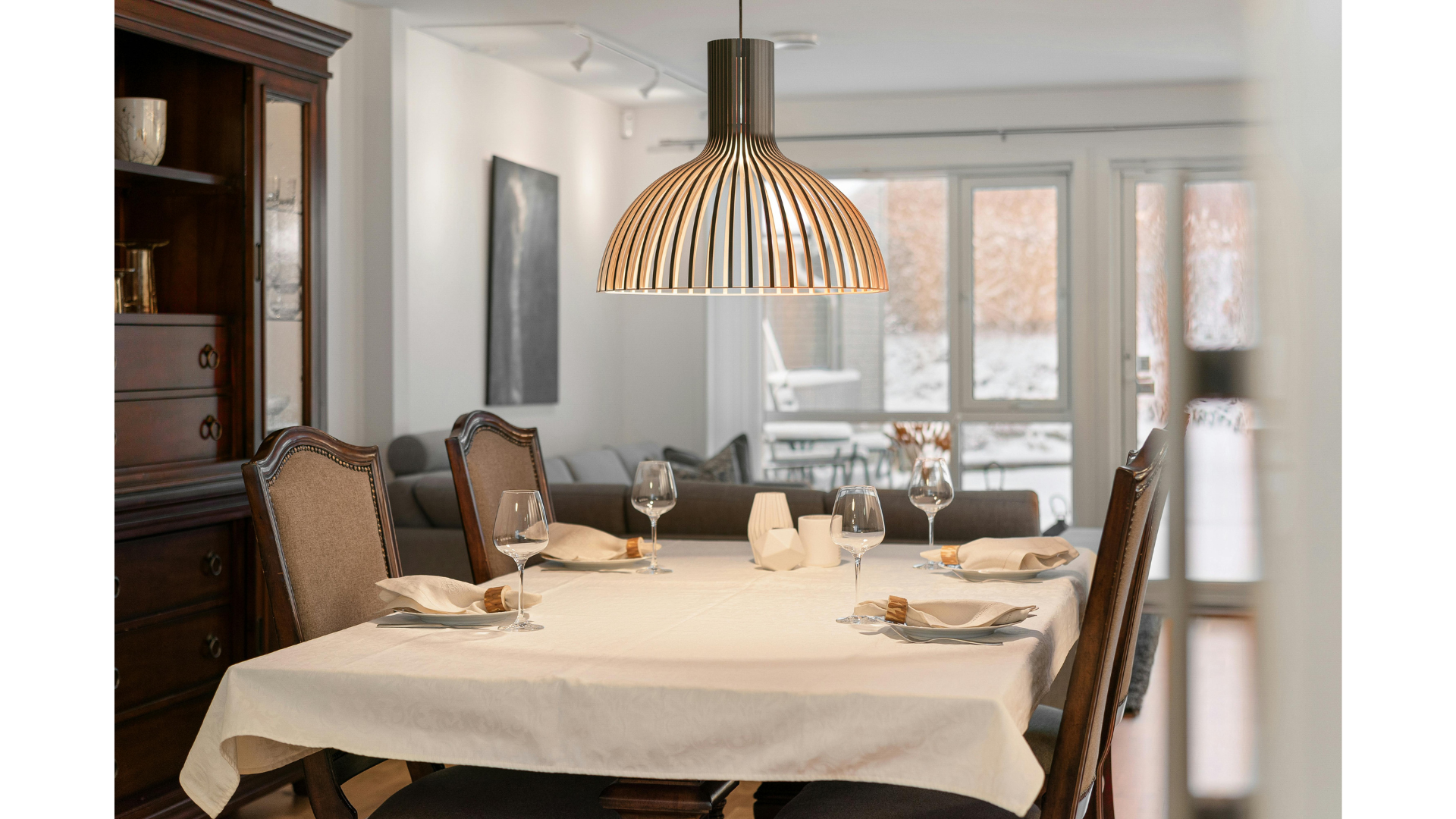 Dining room with a table set for four, featuring white napkins, wine glasses, and small decorative items, illuminated by a large hanging pendant light. In the background, a wooden cabinet and large windows showing a snowy outdoor scene.