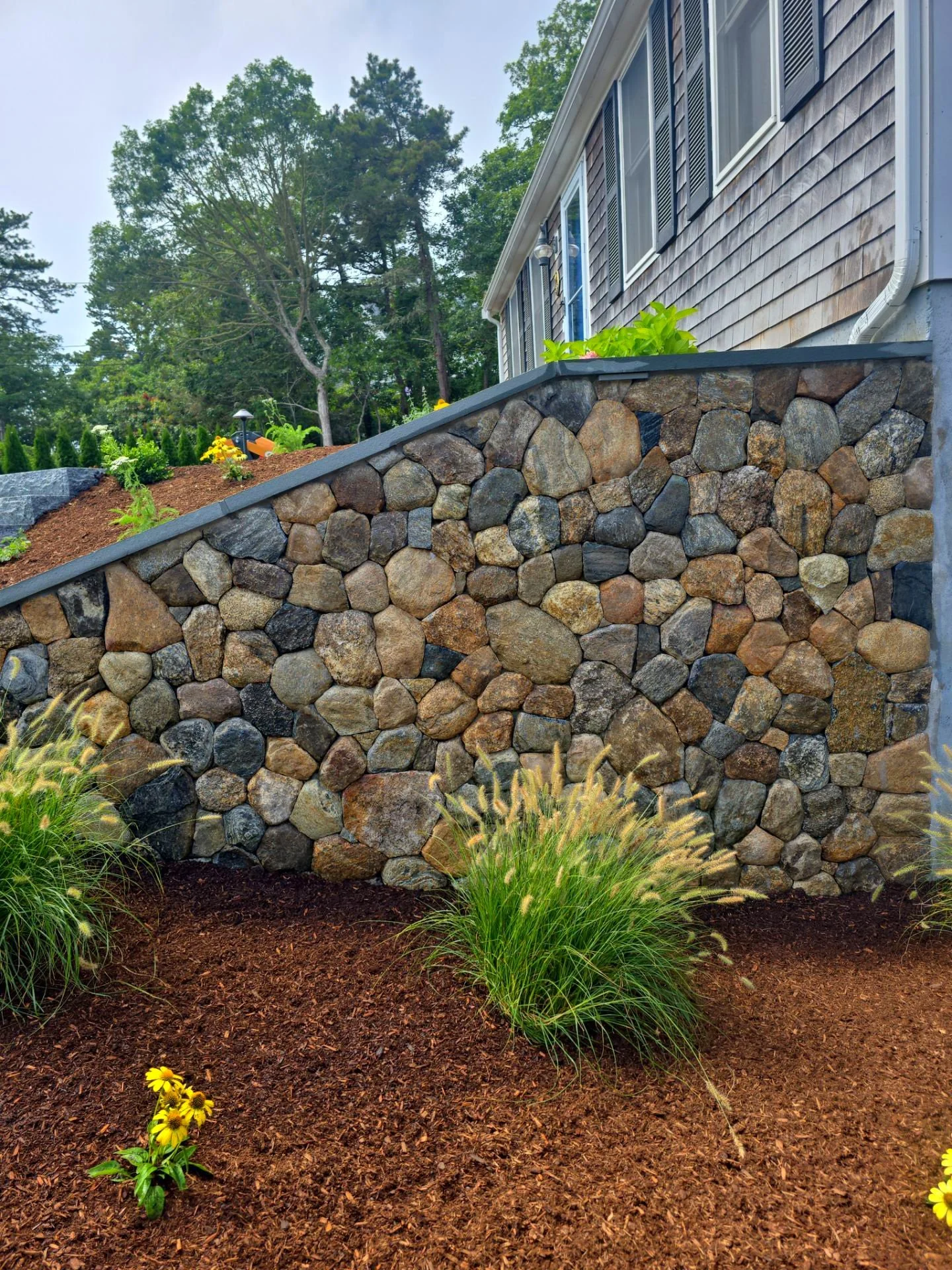 A landscaped yard with a stone retaining wall, a flower bed on top, and some ornamental grasses and yellow flowers in the foreground, next to a house with gray siding and windows.