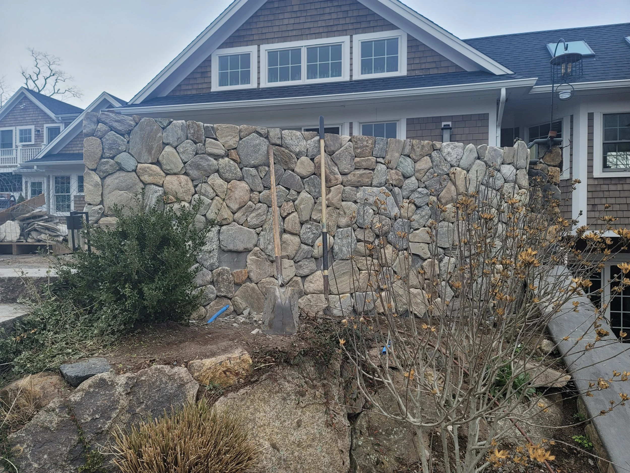 A stone retaining wall in front of a house with a sagging landscape, a bush, a small tree without leaves, gardening tools, and construction materials nearby.