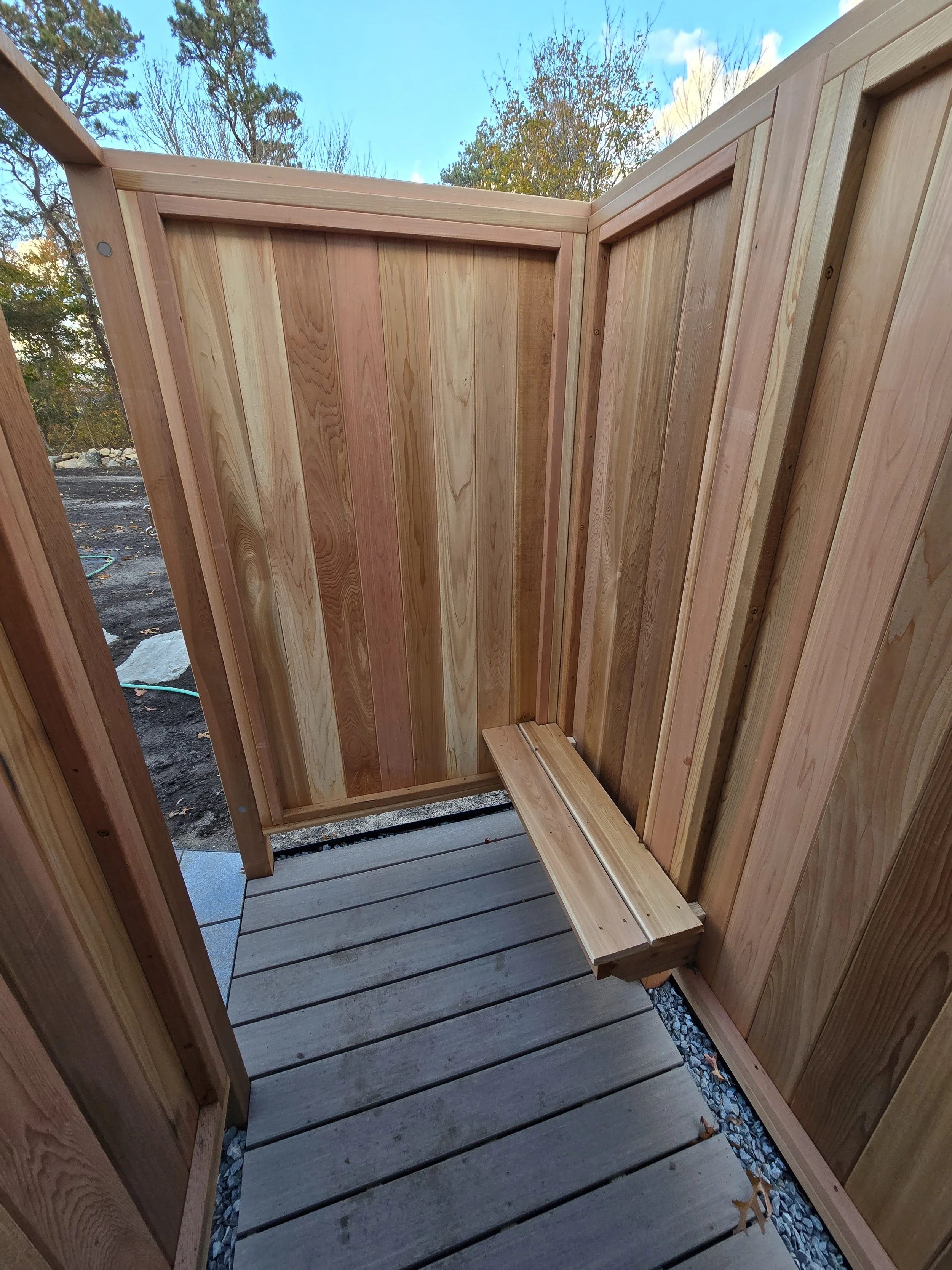 Wooden outdoor shower stall with a small wooden bench, on a deck with a sky and trees in the background.