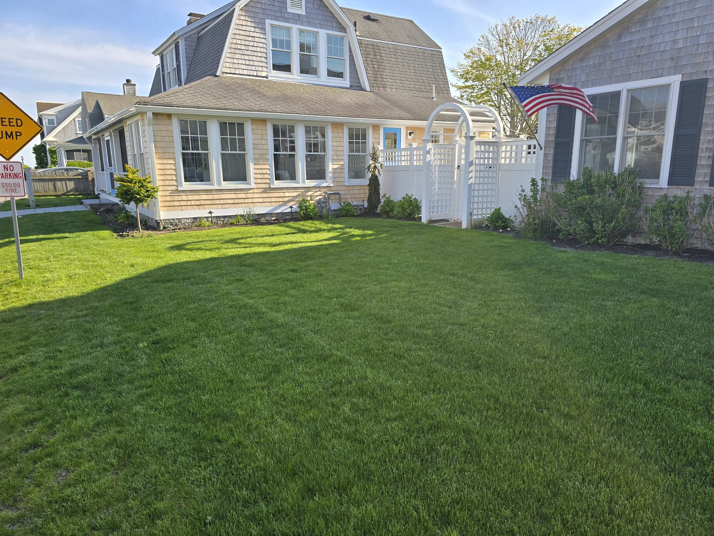 A well-maintained green lawn in front of two houses with a white picket fence and an American flag. A yellow traffic sign partially visible on the left, and a small tree near the house on the left.