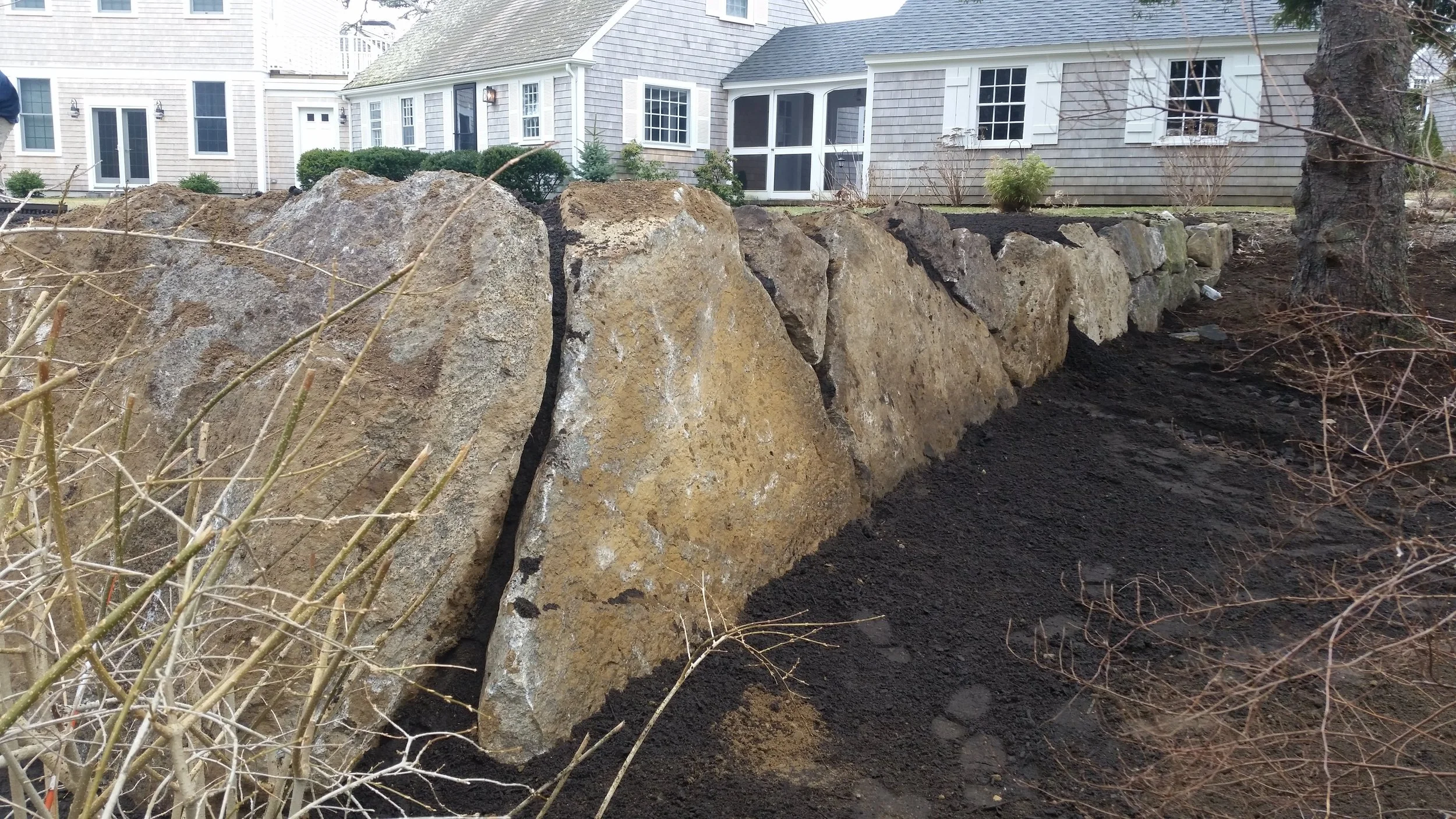 A row of large rocks forming a retaining wall in a garden area, with dark soil and some bare branches in the foreground and houses with white and gray exterior in the background.